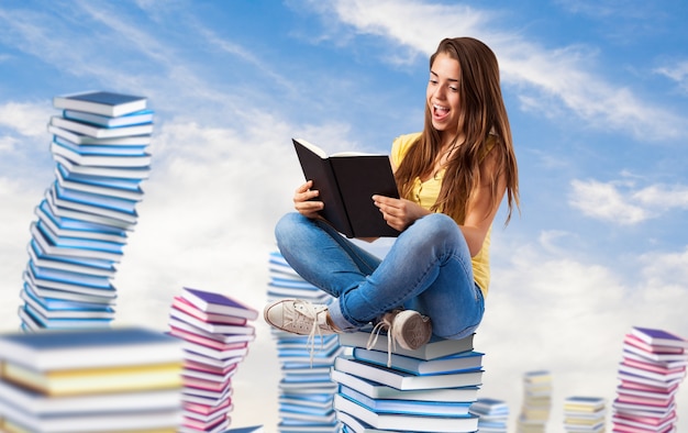 Mujer joven que lee un libro que se sienta en una pila de libros en el cielo