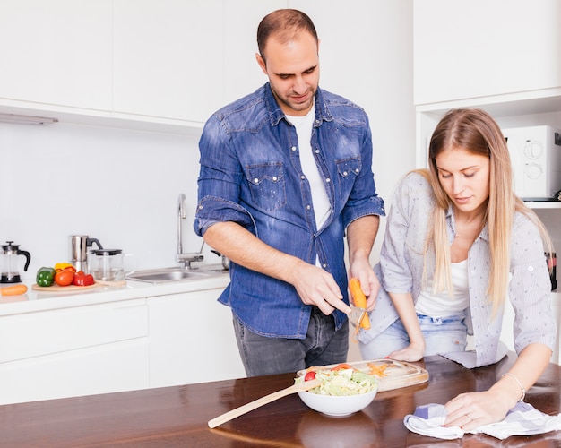 Mujer joven que limpia la mesa con la servilleta y su marido preparando la ensalada en la cocina