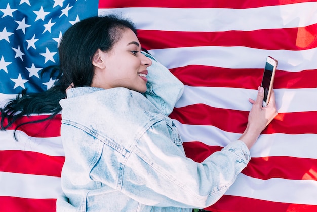 Mujer joven que miente en la bandera americana y que usa smartphone