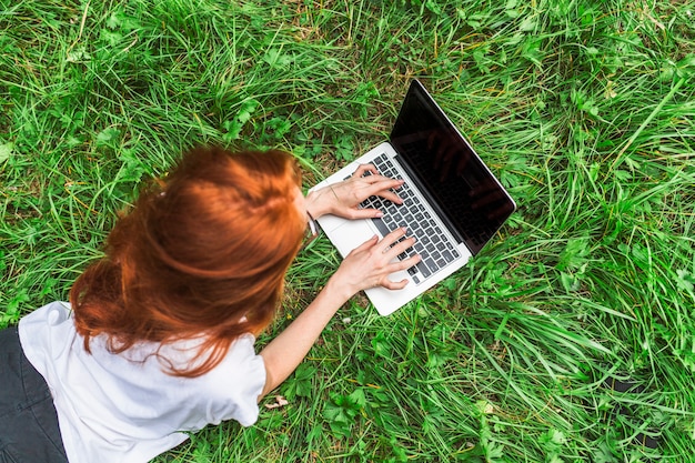 Mujer joven que miente en hierba brillante con la computadora portátil