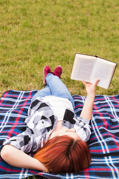 Foto gratuita mujer joven que miente en el libro de lectura de la manta en el parque