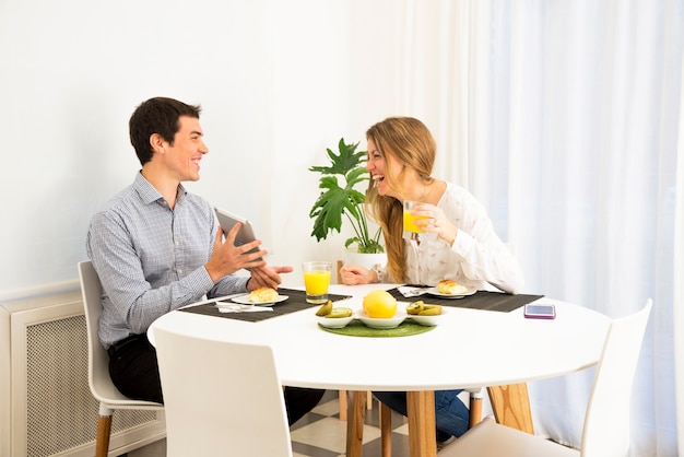 Mujer joven que mira al hombre sonriente que muestra su tableta digital en la mesa de desayuno