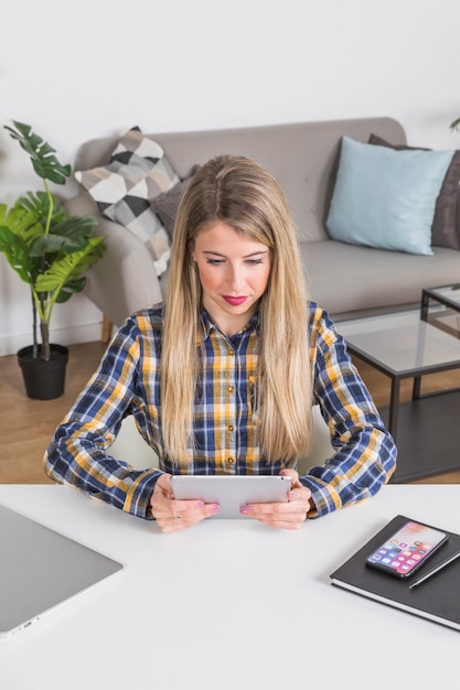 Mujer joven que mira la tableta digital en el escritorio en el interior casero
