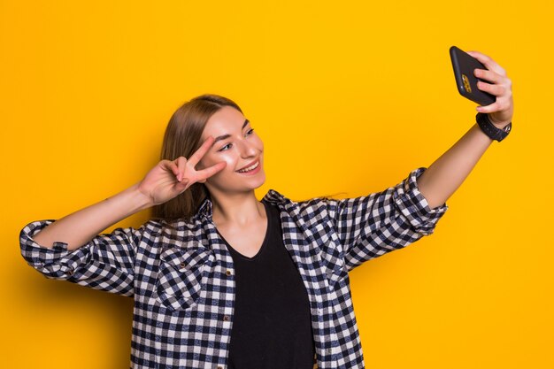Mujer joven que muestra los dedos de la paz y que toma la foto del selfie aislada sobre la pared amarilla