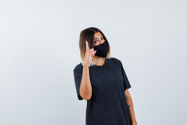 Mujer joven que muestra espera en un gesto de minuto en vestido negro, máscara negra y mirada seria. vista frontal.