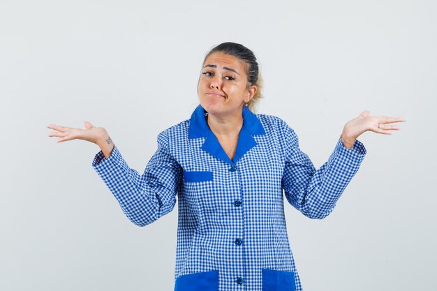 Mujer joven que muestra un gesto de impotencia en camisa de pijama de cuadros azules y luciendo bonita. vista frontal.