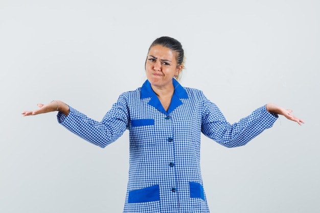 Foto gratuita mujer joven que muestra un gesto de impotencia en camisa de pijama de cuadros azules y mirada incierta. vista frontal.