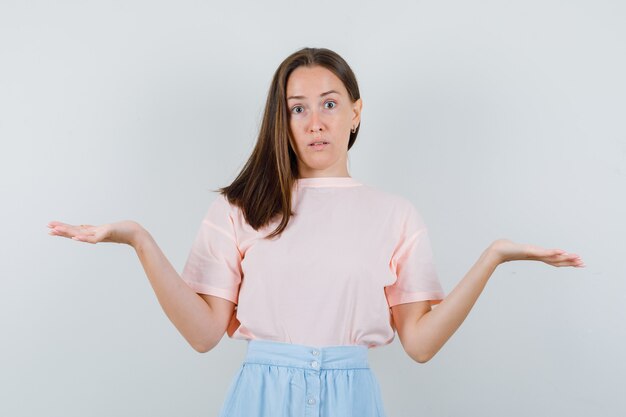 Mujer joven que muestra un gesto de impotencia en camiseta, falda y mirando perplejo. vista frontal.