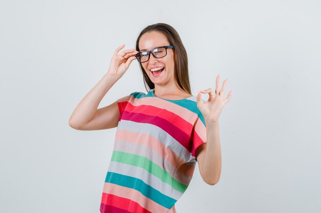 Mujer joven que muestra un gesto de ok con los dedos en las gafas en camiseta y parece alegre. vista frontal.