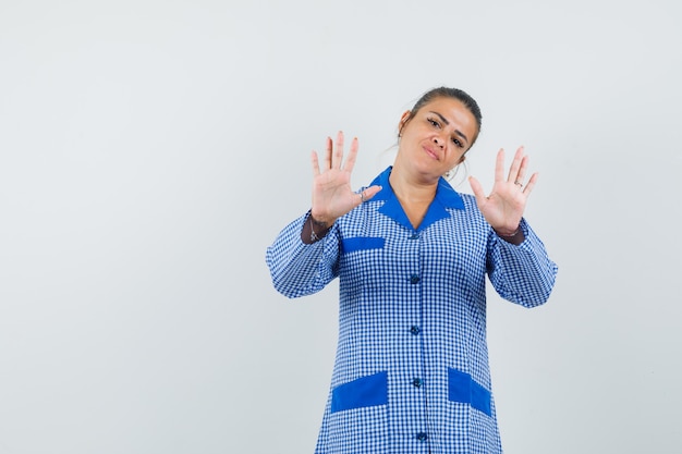 Mujer joven que muestra gesto de restricción en camisa de pijama de cuadros azules y luciendo bonita. vista frontal.