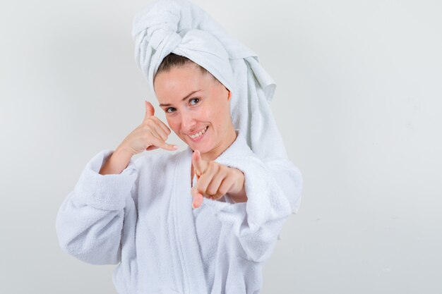 Mujer joven que muestra el gesto del teléfono, apuntando a la cámara en bata de baño blanca, toalla y mirando feliz, vista frontal.