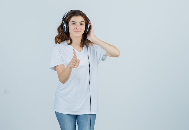 Mujer joven que muestra el pulgar hacia arriba mientras disfruta de la música con auriculares en camiseta blanca, jeans y parece feliz. vista frontal.