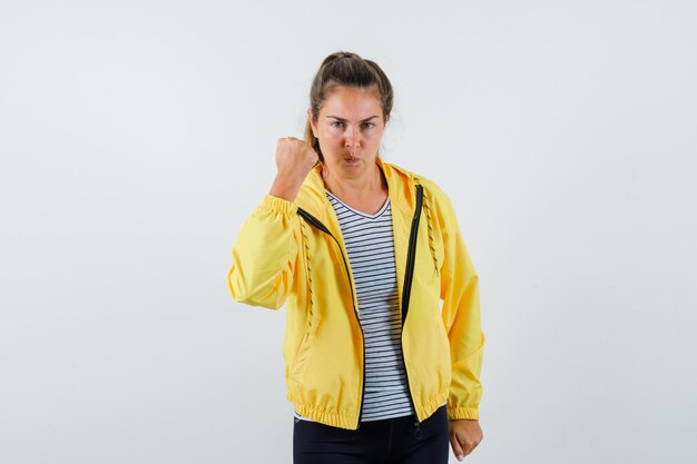 Mujer joven que muestra el puño cerrado en chaqueta, camiseta y parece seguro. vista frontal.