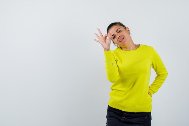 Mujer joven que muestra el signo de ok mientras mantiene la mano detrás de la cintura en un suéter amarillo y pantalón negro y se ve feliz