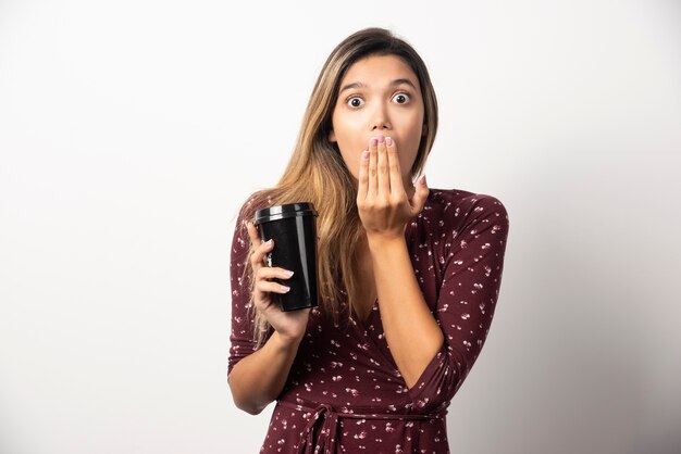 Mujer joven que muestra una taza de bebida en la pared blanca.