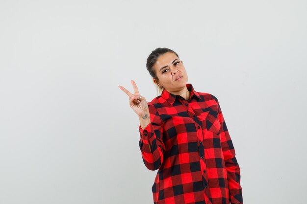 Mujer joven que muestra v-sign en camisa a cuadros y mirando confiado, vista frontal.