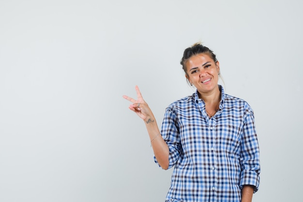 Mujer joven que muestra v-sign en camisa a cuadros y parece alegre.