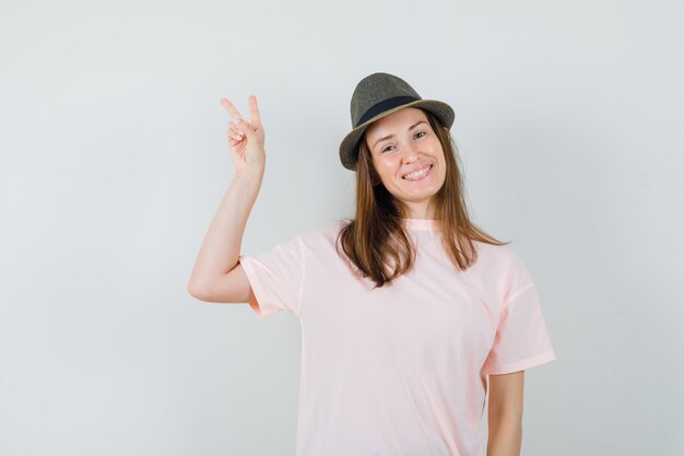 Mujer joven que muestra v-sign en camiseta rosa, sombrero y mirando alegre, vista frontal.