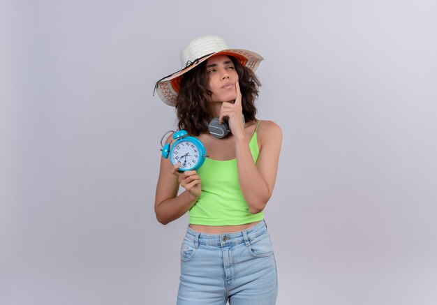 Una mujer joven que piensa con el pelo corto en la parte superior de la cosecha verde con sombrero para el sol manteniendo el dedo índice en la barbilla sosteniendo el reloj de alarma azul sobre un fondo blanco.