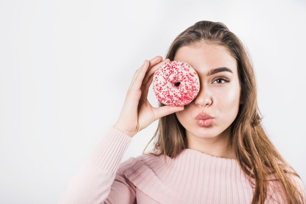 Mujer joven que pone mala cara sus labios que cubren sus ojos con el buñuelo en el contexto blanco