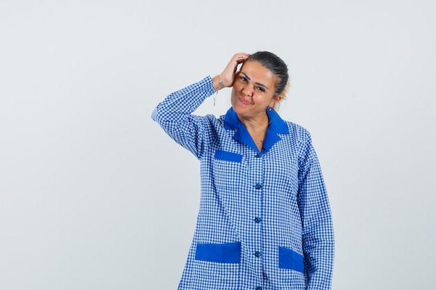 Foto gratuita mujer joven que pone la mano en la cabeza mientras piensa en algo en camisa de pijama de cuadros azules y se ve bonita. vista frontal.