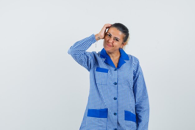 Mujer joven que pone la mano en la cabeza mientras piensa en algo en camisa de pijama de cuadros azules y se ve bonita. vista frontal.