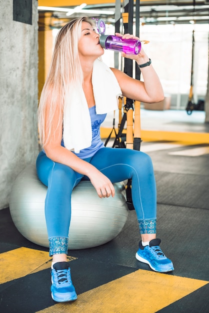 Mujer joven que se sienta en el agua potable de la bola de la aptitud en gimnasio