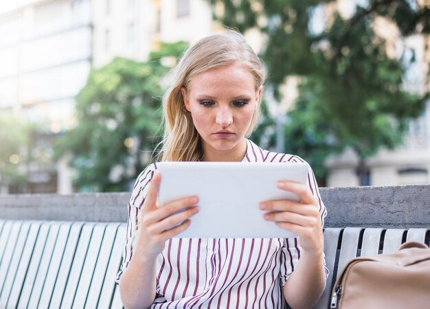 Mujer joven que se sienta en el banco que sostiene la tableta digital