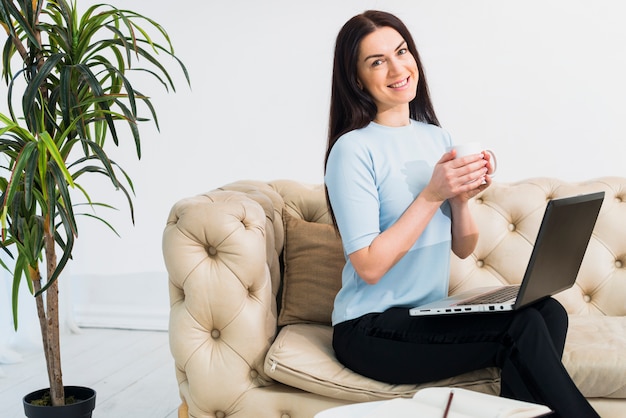 Mujer joven que se sienta con café y la computadora portátil en el sofá
