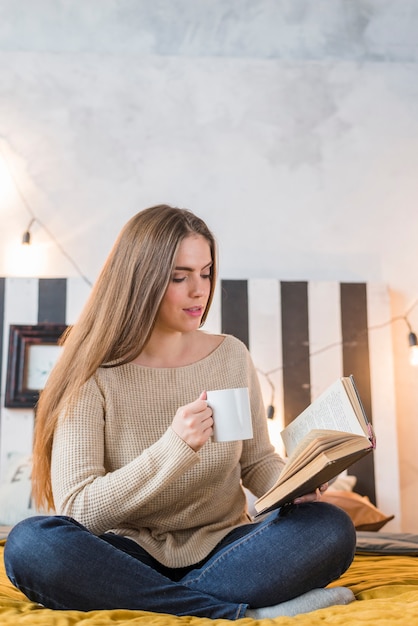 Foto gratuita mujer joven que se sienta en la cama que sostiene la taza de café en el libro de lectura de la mano