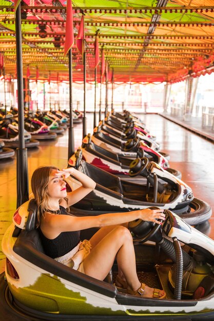 Mujer joven que se sienta en el coche de parachoques que protege sus ojos en el parque de atracciones