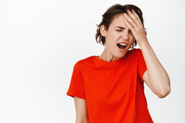 Mujer joven que siente dolor, dolor de cabeza, toca la frente y hace muecas por la incomodidad, se siente enferma, de pie en camiseta en blanco