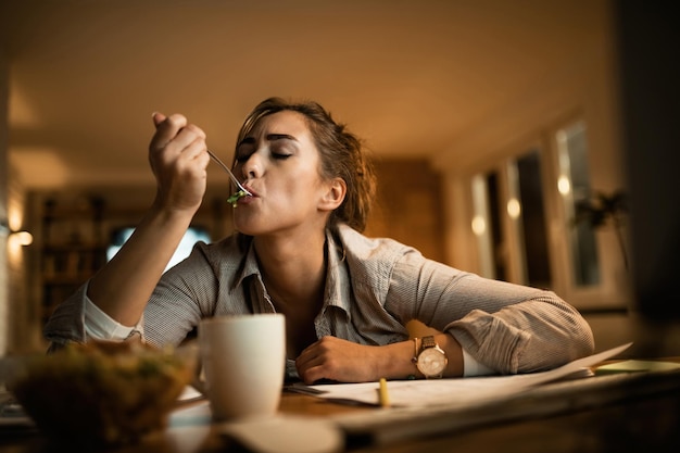 Mujer joven que siente hambre y come ensalada mientras estudia por la noche en casa