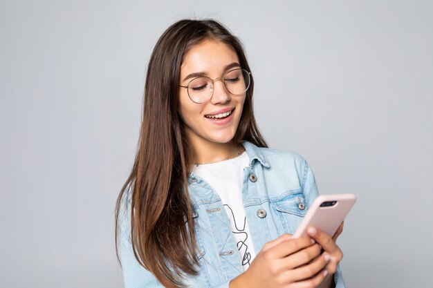 Mujer joven que sonríe y que manda un SMS en su teléfono móvil, aislado sobre la pared blanca.