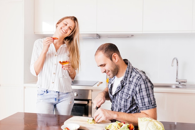 Foto gratuita mujer joven que sostiene la copa disponible en la mano que mira a su marido que corta la verdura en la cocina