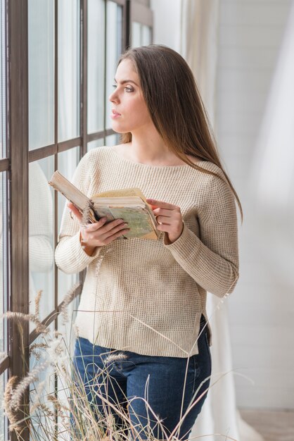 Mujer joven que sostiene el libro en la mano mirando a través de la ventana