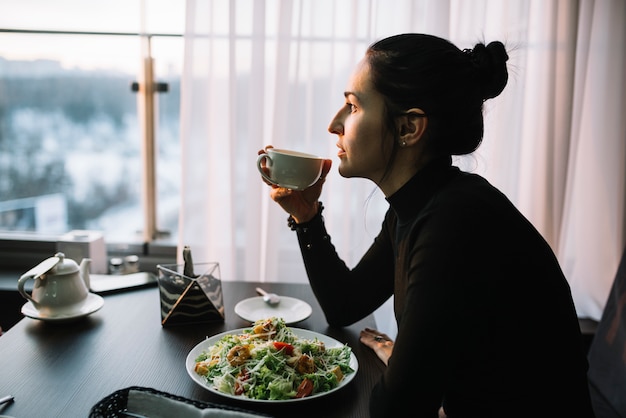 Foto gratuita mujer joven que sostiene la taza de bebida en la mesa con ensalada cerca de la ventana