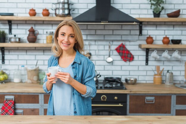 Mujer joven que sostiene la taza de café en la mano que se coloca en la cocina