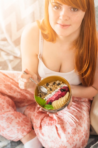 Mujer joven que sostiene el tazón de granola de avena casera con arándanos; Rodajas de frambuesa y plátano