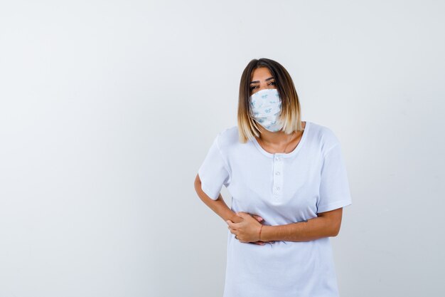 Mujer joven que sufre de dolor de estómago en camiseta, máscara y mal aspecto. vista frontal.