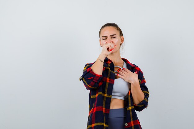 Mujer joven que sufre de tos en la parte superior de la cosecha, camisa a cuadros, pantalones y con aspecto de malestar. vista frontal.