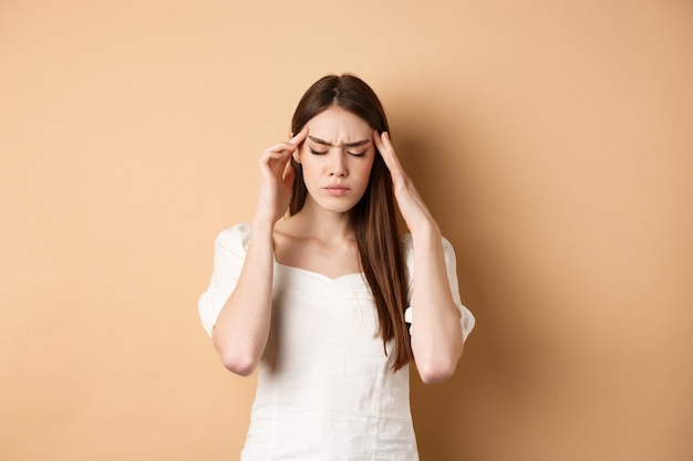 Mujer joven que tiene dolor de cabeza tocando las sienes de la cabeza con los ojos cerrados y la cara tensa de pie ...