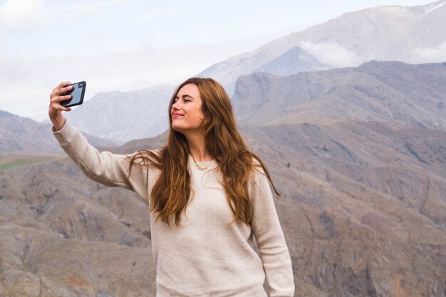 Mujer joven que toma autofotos delante del paisaje de montaña