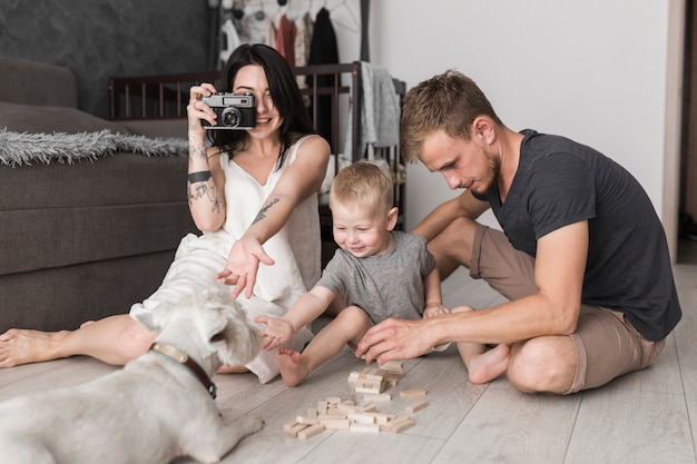 Mujer joven que toma la foto del perro con la cámara sentada cerca de su hijo y su esposo jugando juntos