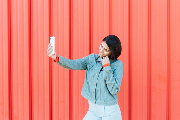 Mujer joven que toma el selfie en el teléfono móvil que se opone al fondo metálico rojo