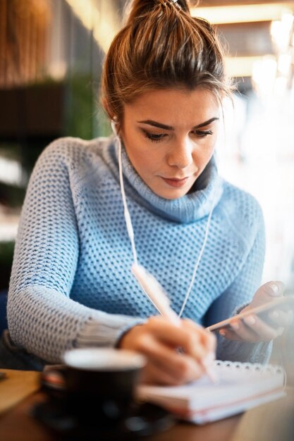 Mujer joven que trabaja en un café y escribe notas en un planificador semanal mientras usa el teléfono móvil y escucha música con auriculares