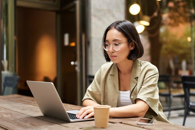 Mujer joven que trabaja en un café usando una computadora portátil y bebiendo café estudiante asiática con semental de computadora