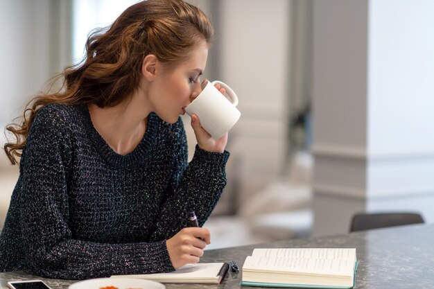 Mujer joven que trabaja en casa usando el Bloc de notas en la cocina. Ella está tomando café. Ideas para negocios. Estudiar y trabajar desde casa.