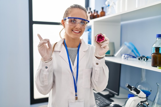 Mujer joven que trabaja en el laboratorio científico sosteniendo geoda sonriendo feliz señalando con la mano y el dedo a un lado