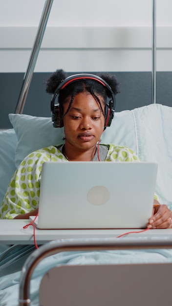 Foto gratuita mujer joven que usa auriculares y una computadora portátil para divertirse en la sala del hospital mientras espera para curar la enfermedad. paciente enfermo que usa auriculares para escuchar música y se relaja en la cama con un monitor de frecuencia cardíaca